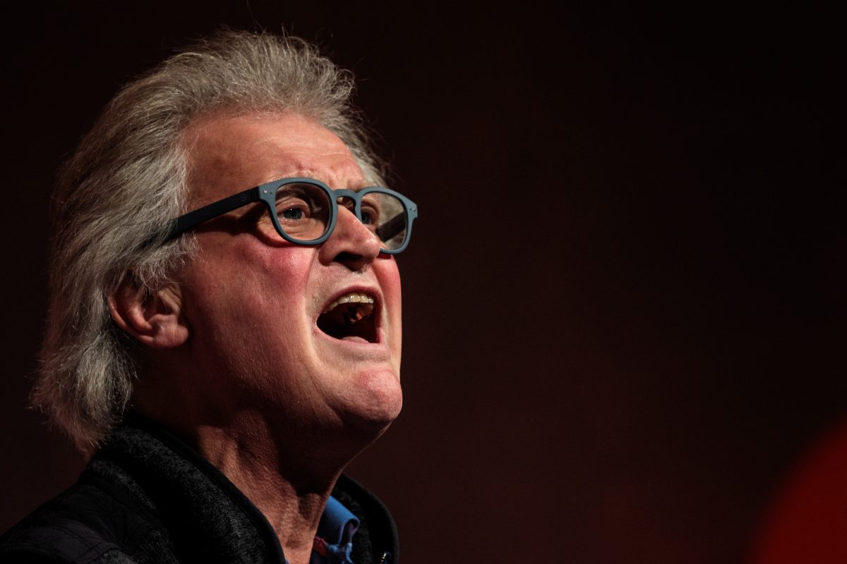 business Older man with grey hair and blue glasses speaking passionately, mouth open, against a dark background.