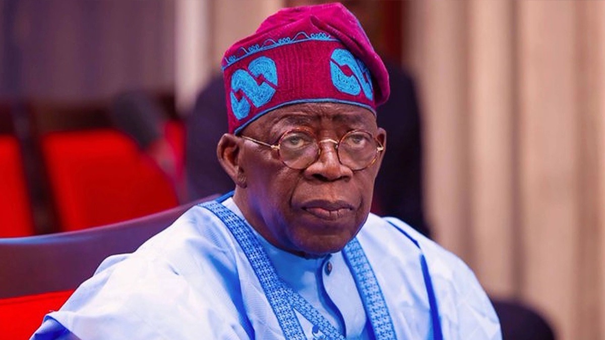 Tinubu - THEWILL NEWS MEDIA Portrait of an older African man wearing a blue traditional outfit and a red patterned cap, with glasses, seated indoors.